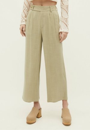 Pantalon beige taille haute à jambes larges associé à des sabots à plateforme beiges et un haut blanc à manches longues en crochet avec abdomen découvert.