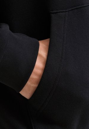 Čierna mikina, zameraná na detail švu a vrecka, pričom je čiastočne vidieť ruku spočívajúcu vo vrecku. Mäkká textúra.