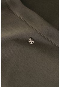 Primo piano di un tessuto verde oliva con un piccolo medaglione metallico rotondo con un design a motivi, che mette in evidenza una consistenza morbida e una leggera lucentezza.