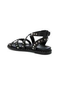 Sandal de cuero negro con tiras entrelazadas, detalles de tachuelas y suela plana. Presenta tachuelas metálicas a lo largo de las tiras y una plantilla texturizada.