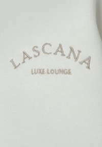 Witte lounge top met geborduurde tekst "LASCANA LUXE LOUNGE" in beige draad, zachte stof met een gladde textuur.