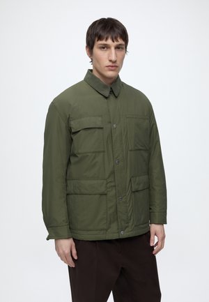 Mann trägt eine olivgrüne Jacke mit vier Vorderentaschen und dunkle Hosen, steht vor einem einfarbigen weißen Hintergrund.