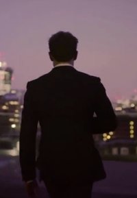 Una persona in un elegante completo nero con una camicia bianca si allontana, silhouettata contro lo skyline di una città al crepuscolo. Sono visibili edifici illuminati.