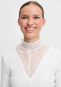 Hvid langærmet top med en høj blondekrave, der har indviklede mønstre og bølgede kanter. Blød, ribbet tekstur fremhæver designet.