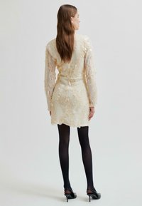 Kobieta stoi tyłem w kremowej, cekinowej mini sukience, czarnych rajstopach i czarnych butach na obcasie, na tle jednolitego, jasnego tła.