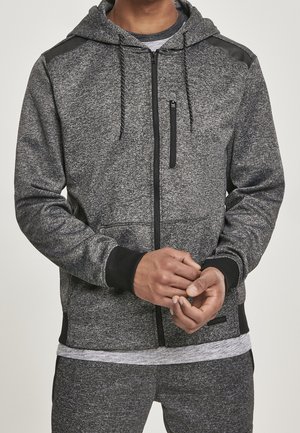Homme portant un sweat à capuche gris zippé devant avec un galon noir et un pantalon gris assorti, les mains jointes devant.