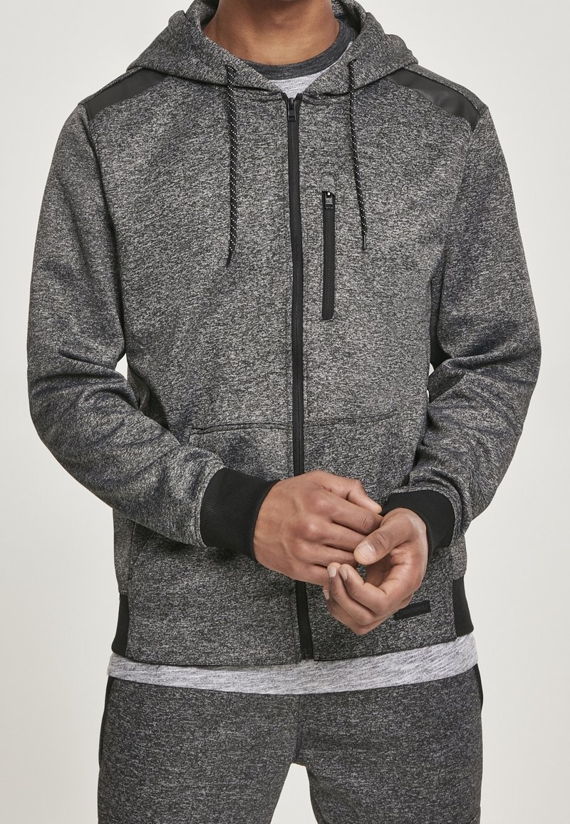 Homme portant un sweat à capuche gris zippé devant avec un galon noir et un pantalon gris assorti, les mains jointes devant.