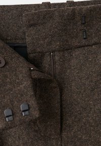 Il tessuto marrone testurizzato presenta una chiusura con zip, accenti in bottone e passanti per cintura rettangolari. Il design enfatizza una vestibilità su misura.