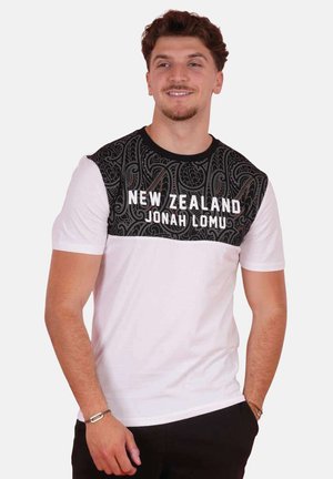 Homme portant un t-shirt blanc et noir avec le texte "New Zealand Jonah Lomu", debout avec une main dans la poche, souriant légèrement, regardant vers la droite.