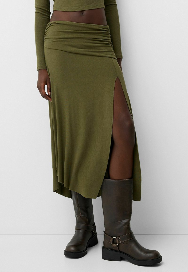 Jupe olive, longueur genou avec une fente haute, taille froncée et texture douce, associée à des bottes marron jusqu'au genou et un haut ajusté à manches longues.