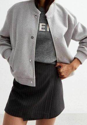 Personne portant une veste boutonnée gris clair, un pull gris avec un texte partiel et une jupe sombre à fines rayures, debout avec une main dans la poche.