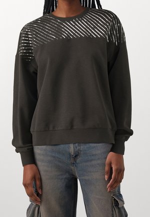 Osoba ubrana w czarną bluzę z ukośnymi srebrnymi paskami na ramionach oraz wyblakłe niebieskie spodnie typu cargo z kieszeniami.