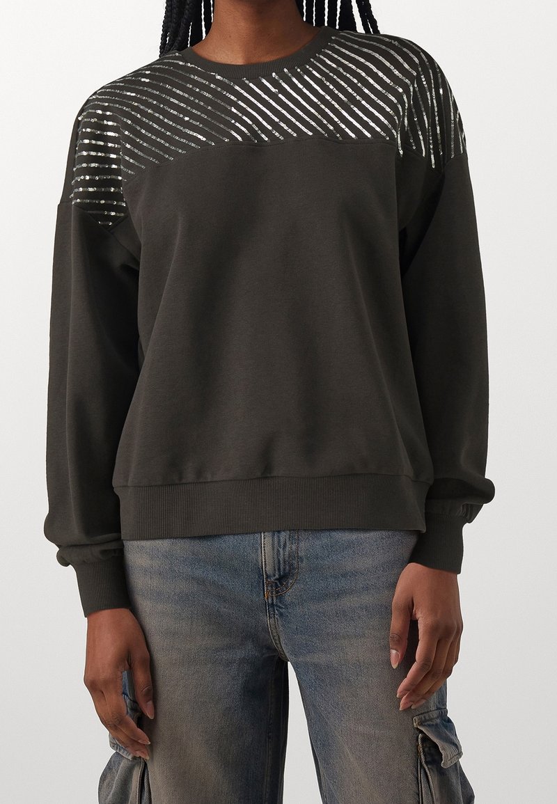 Personne portant un sweat-shirt noir avec des bandes diagonales argentées sur les épaules et un jean cargo bleu délavé avec des poches.