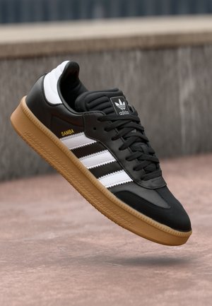 Czarny sneaker Adidas Samba z białymi paskami, gumową podeszwą i żółtym napisem "SAMBA", unoszący się nad teksturowaną powierzchnią ziemi.