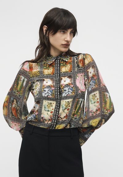 Γυναίκα που φοράει κουμπωτό, μακρυμάνικο μπλουζάκι με πολύχρωμες εκτυπώσεις καρτών ταρό, συνδυασμένο με μαύρο παντελόνι ψηλόμεσο.