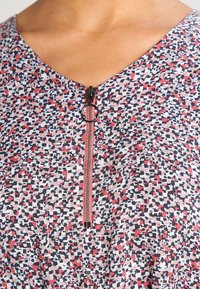 Haut multicolore présentant un motif vibrant en rouge, noir et blanc. Il a une courte fermeture éclair avec un tirette circulaire au niveau du col.