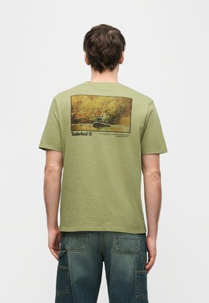 Homme portant un t-shirt Timberland vert olive avec une photo imprimée dans le dos montrant deux personnes pêchant sur un bateau dans une forêt d'automne.