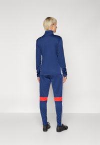 Navy tracksuit with high collar and long sleeves, featuring white side stripes and red accents on pants. Smooth, athletic material.