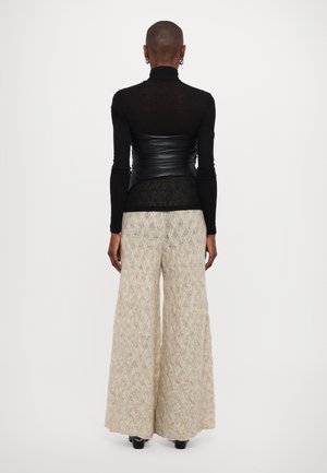 Personne aux cheveux courts portant un haut noir à manches longues, un corset en cuir noir, un pantalon large beige texturé et des chaussures noires, debout de dos.