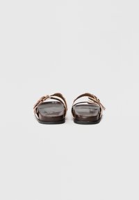 Καφέ επίπεδες σάντες με μαλακή δερμάτινη σόλα, φαρδιές λωρίδες και διακοσμητικά στοιχεία σε ανοιχτό ροζ. Ο σχεδιασμός περιλαμβάνει μια λεπτή υφή.