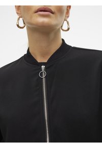 Veste bomber noire avec col côtelé et fermeture éclair argentée. Gros plan sur la texture du tissu et le détail de la tirette de fermeture éclair circulaire.