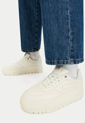 Zapatillas blancas con plataforma, combinadas con calcetines blancos y vaqueros azules, mostradas desde ángulos lateral y frontal sobre un fondo blanco.