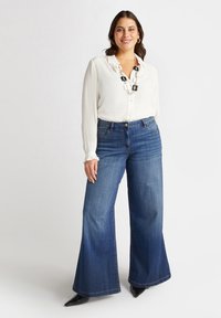 Biała bluzka z długim rękawem i falbaniastym kołnierzykiem, zestawiona z niebieskimi spodniami o szerokich nogawkach. Spodnie wyróżniają się klasyczną teksturą denimu i subtelnym przeszyciem.