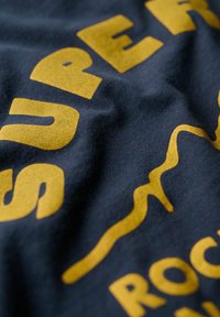Tissu foncé avec des lettres jaunes en gras affichant partiellement "SURE" et une ligne ondulée, probablement une partie d'un logo ou d'un design textuel sur un vêtement.