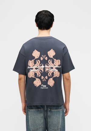 Osoba ubrana w ciemnoszarą koszulkę z różowym, abstrakcyjnym kwiatowym wzorem oraz białym napisem na plecach, w parze z niebieskimi dżinsami.