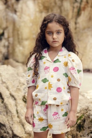 Ragazza giovane con capelli lunghi e ricci che indossa un completo bianco composto da maglietta e pantaloncini abbinati, decorati con pianeti colorati, stelle e UFO.