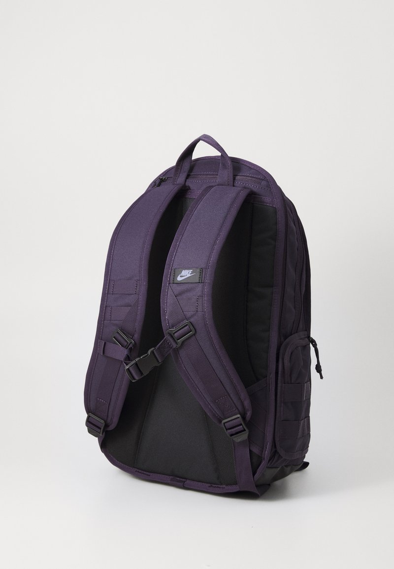 Sac à dos en nylon violet avec des bretelles rembourrées, des compartiments zippés et une poche latérale. Présente un petit patch logo à l'avant.