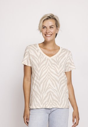 Smiling woman with short blonde hair wearing a beige and white zebra-patterned V-neck T-shirt and light blue jeans against a plain background.