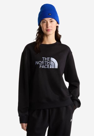 Nainen, joka pukeutunut mustaan The North Face -collegepaitaan ja housuihin, sekä kirkkaan siniseen neulottuun pipoon, seisoo yksivärisen valkoisen taustan edessä.