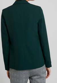 Žena v tmavě zeleném přizpůsobeném saku a šedých kostkovaných kalhotách, zobrazená ze zad na světlém jednobarevném pozadí.