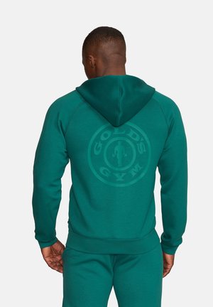 Mężczyzna w zielonkawym bluzy z kapturem i dopasowanych spodniach, pokazujący logo Gold's Gym nadrukowane na plecach bluzy.