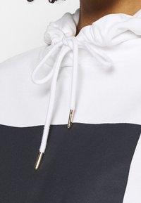 Sudadera con la mitad superior blanca y la mitad inferior gris oscuro. Capucha con cordones y extremos de metal en tono dorado; textura suave.