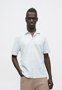 Lyseblå kortermet poloskjorte med strukturert mønster, med knappnedkrage og rett nederkant, kombinert med beige bukser.