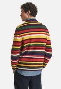 Multikleurige gestreepte trui met een mix van rood, groen, geel en wit. Geribbelde kraag en manchetten, met een dennenhemdzoom zichtbaar eronder.
