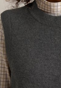 Dunkelgrauer ärmelloser Strickpullover mit einer weichen, strukturierten Oberflächenbeschaffenheit, der über einem hellbeigen karierten Hemd mit Kragen getragen wird.