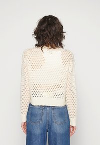 Pull cropped beige clair avec un design en maille lâche, manches longues et ourlet côtelé. Porté avec un jean bleu taille haute.
