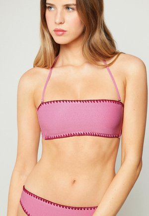 Jeune femme portant un haut de bikini rose sans bretelles avec des surpiqûres rose foncé le long des bords, tournée légèrement de côté sur un fond uni.