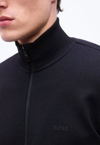 Κοντινό πλάνο ενός ατόμου που φοράει μαύρο μπουφάν με φερμουάρ, ριμπ κολάρο και λογότυπο "BOSS" στο στήθος.