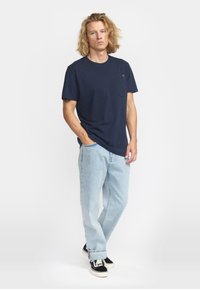 Camiseta de algodón azul marino con un bolsillo en el frente y un pequeño logo colorido, combinada con jeans azul claro y zapatillas negras.