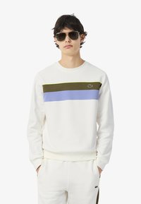 Sudadera blanca con rayas horizontales en oliva, púrpura y verde. Presenta un cuello redondo y un pequeño logo en el pecho. Se lleva con gafas de sol.