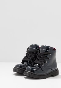 Černé lakované kotníkové boty s černými šňůrkami, stříbrnými očky a červeným akcentem na kotníku. Mají texturovanou gumovou podrážku.