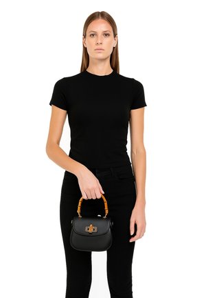 Vrouw in een nauwsluitend zwart T-shirt en broek, met een kleine zwarte handtas met een bruine handgreep in bamboestijl voor zich.
