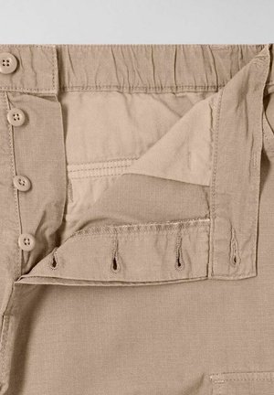 Pantaloni beige con chiusura a bottone, vita elastica e spacco laterale. Il tessuto ha una texture liscia e dettagli di cucitura sottili.