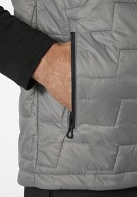 Graue isolierte Weste mit einem gesteppten Muster, ausgestattet mit einer seitlichen Reißverschlusstasche und einem schwarzen Langarmshirt darunter. Glatte Textur.