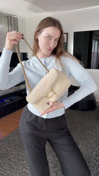 Borsa a mano in pelliccia sintetica beige con chiusura circolare dorata, dotata di tracolla intrecciata in catena dorata. La borsa ha una texture morbida e un design con patta.