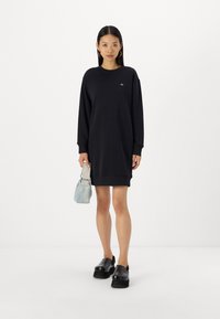 Svart sweatshirtkjole med lange ermer, ribbede mansjetter og rund halsutskjæring. Kombinert med en lys blå håndveske og svarte platåsandaler.